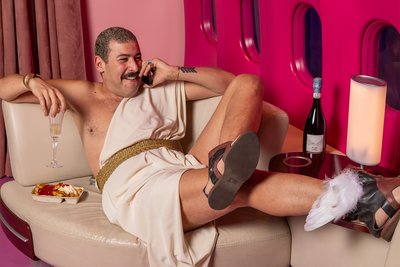 Das Farbfoto zeigt einen Mann mit Schnurrbart. Er liegt entspannt in einer luxuriösen Umgebung, die an das Innere eines Flugzeugs erinnert. Er trägt eine Art Toga mit goldenem Gürtel, an den Füßen Sandalen mit Flügeln. In einer Hand hält er ein Glas Sekt, in der anderen ein Telefon. Neben ihm stehen eine Pappschale mit Pommes Frites. Auf einem Sofa ihm gegenüber liegt ein Helm mit Flügeln. Die Szenerie ist in kräftigem Pink gehalten.