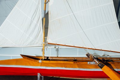 Ein langes Sportsegelboot hängt über dem Holzboden. Es hat drei aufgespannte weiße Segel und einen rotbraunen Rumpf. Im Hintergrund ragt ein weiteres Segel aus weißen, horizontal gespannten Wellenbändern hervor.