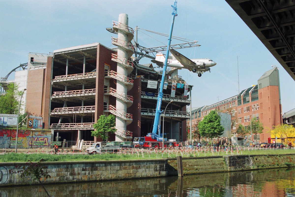 Rohbau eines vierstöckigen Gebäudes. An einer Stahlkonstruktion auf dem Dach hängt ein Flugzeug. Vor dem Gebäude steht ein blauer Kran.