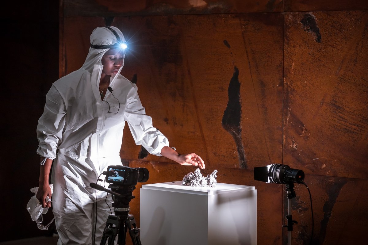 Eine Frau mit dunkler Hautfarbe, in einen weißen Schutzanzug mit Stirnlampe gekleidet, berührt mit einer Hand einen Haufen Staub, der auf einem Sockel liegt. Im Hintergrund ist eine braune Metallwand zu sehen.