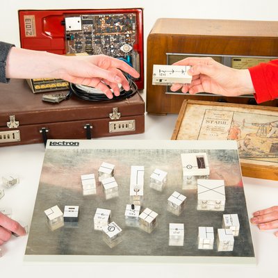 Mehrere historische und elektronische Geräte stehen auf einem Tisch, darunter ein Radio, ein geöffnetes Elektronikgerät und ein Experimentierkasten. Zwei Personen reichen sich ein elektronisches Bauteil über einer Fläche mit modularen Steckelementen des Lernsystems „Lectron“.