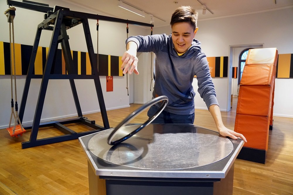 Eine Person stützt sich auf einen Tisch mit metallischer und leicht nach innen gewölbter Oberfläche. Sie dreht mit dem freien Arm einen Ring an, dessen Durchmesser ungefähr 30 cm beträgt.