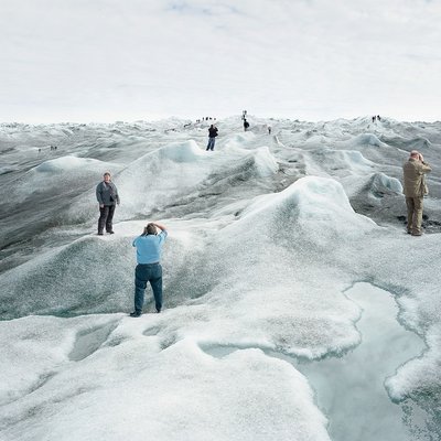 Eine Gruppe von Touristinnen und Touristen steht auf einer schmelzenden Eislandschaft und posiert an verschiedenen Punkten des Bildes für Fotos. An vielen Stellen der Eislandschaft hat sich schwarzer Ruß abgesetzt, das Eis ist dort sehr dunkel. Im Vordergrund ist eine große Pfütze aus Schmelzwasser.