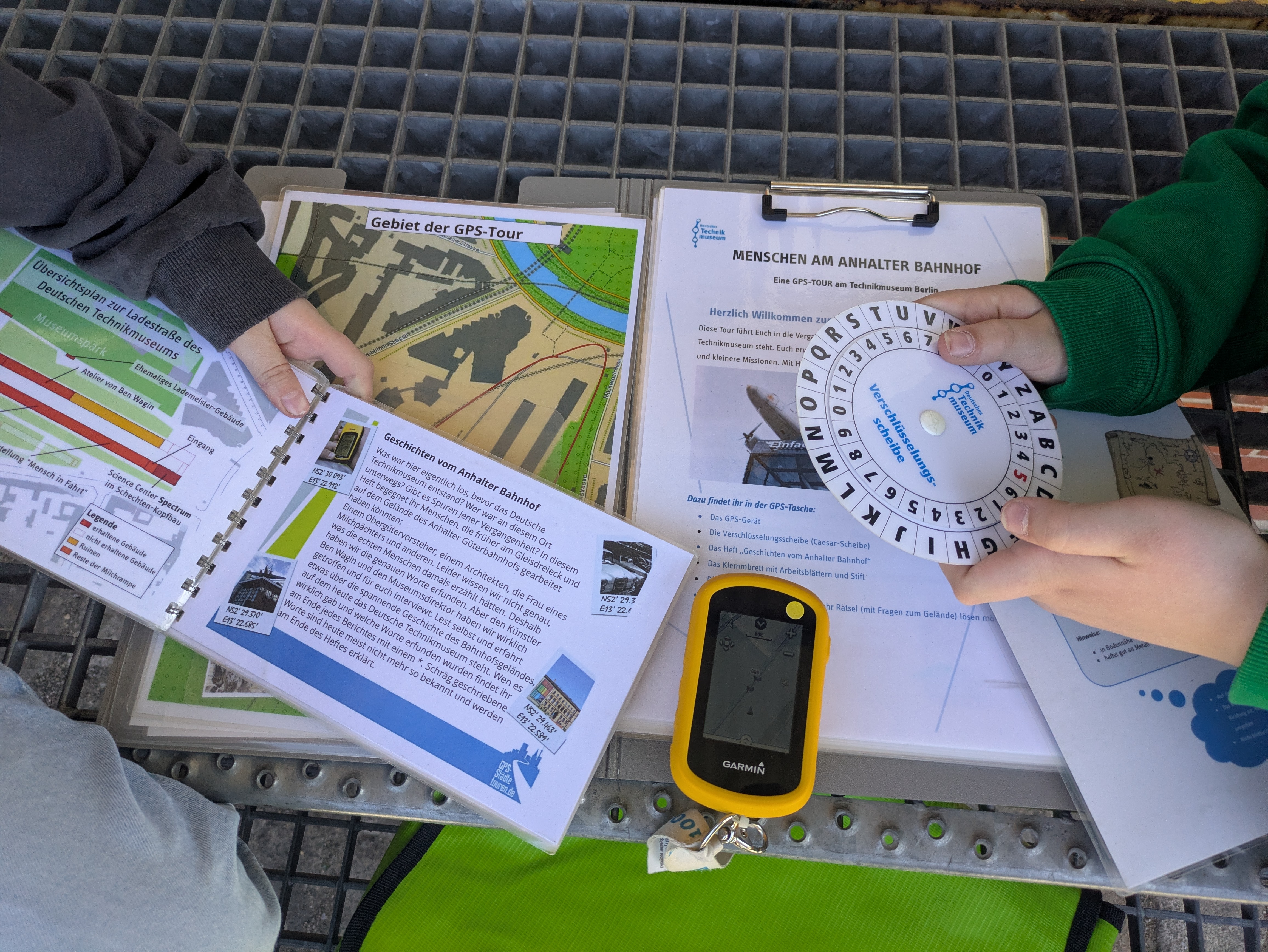 Man sieht die Materialien für die GPS-Tour: Ein GPS-Gerät, eine Verschlüsselungsscheibe und zwei Informationshefte.