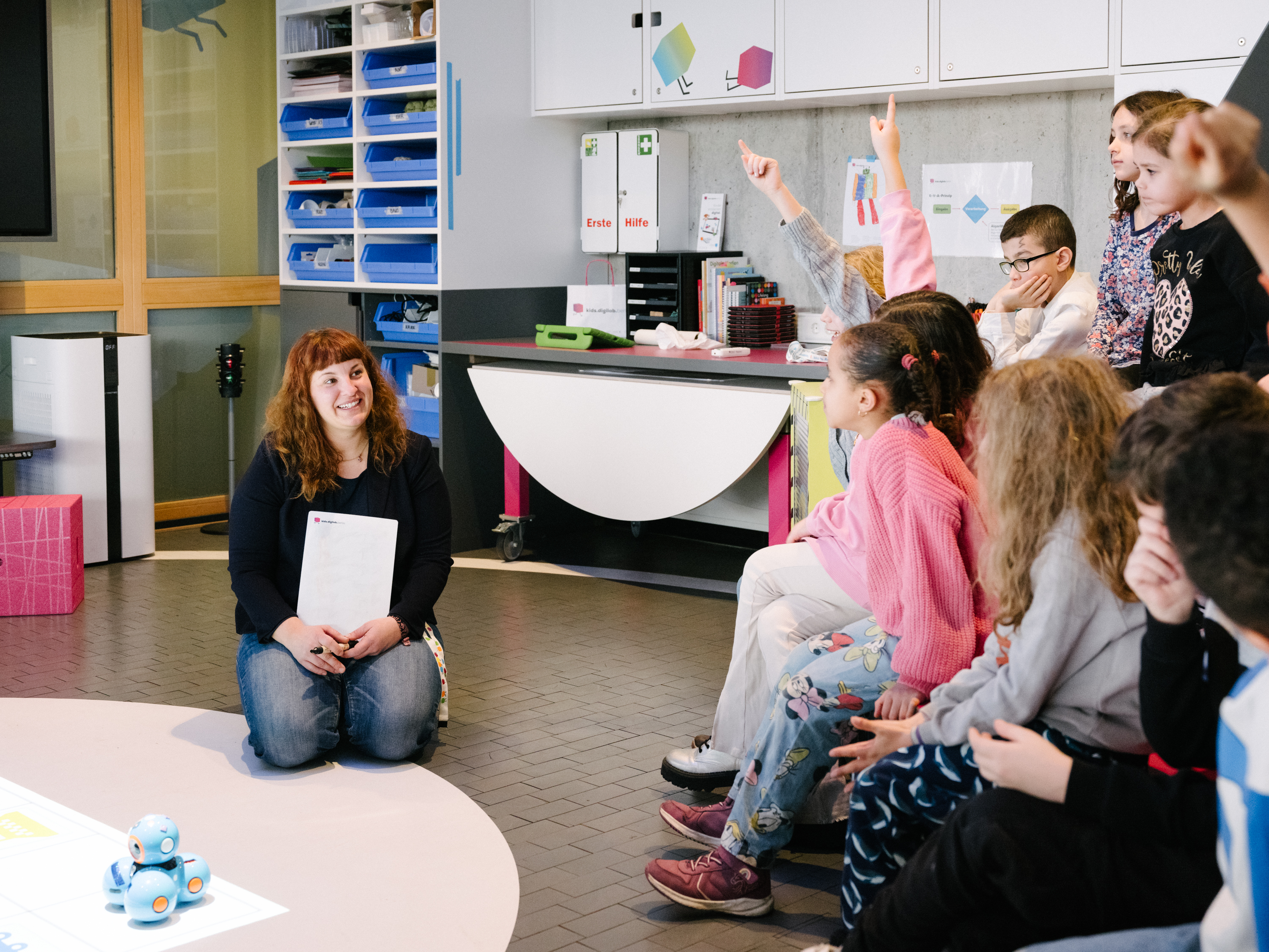 Eine Gruppe von Kindern sitrzt in einem bunten Raum, neben ihnen kniet eine Frau, die etwas erklärt.