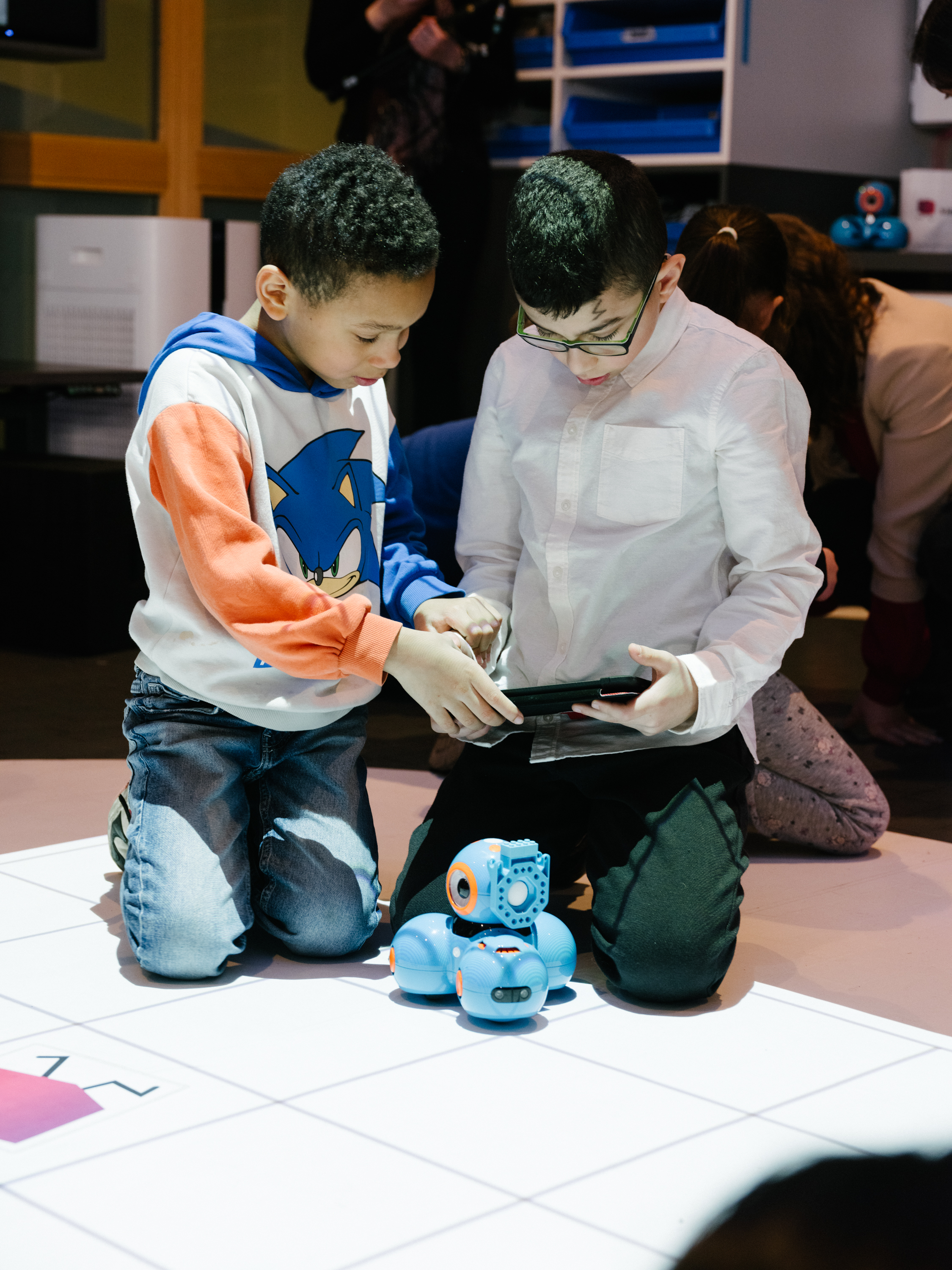Zwei Jungen knien neben einem kleinen blauen Roboter und unterhalten sich.