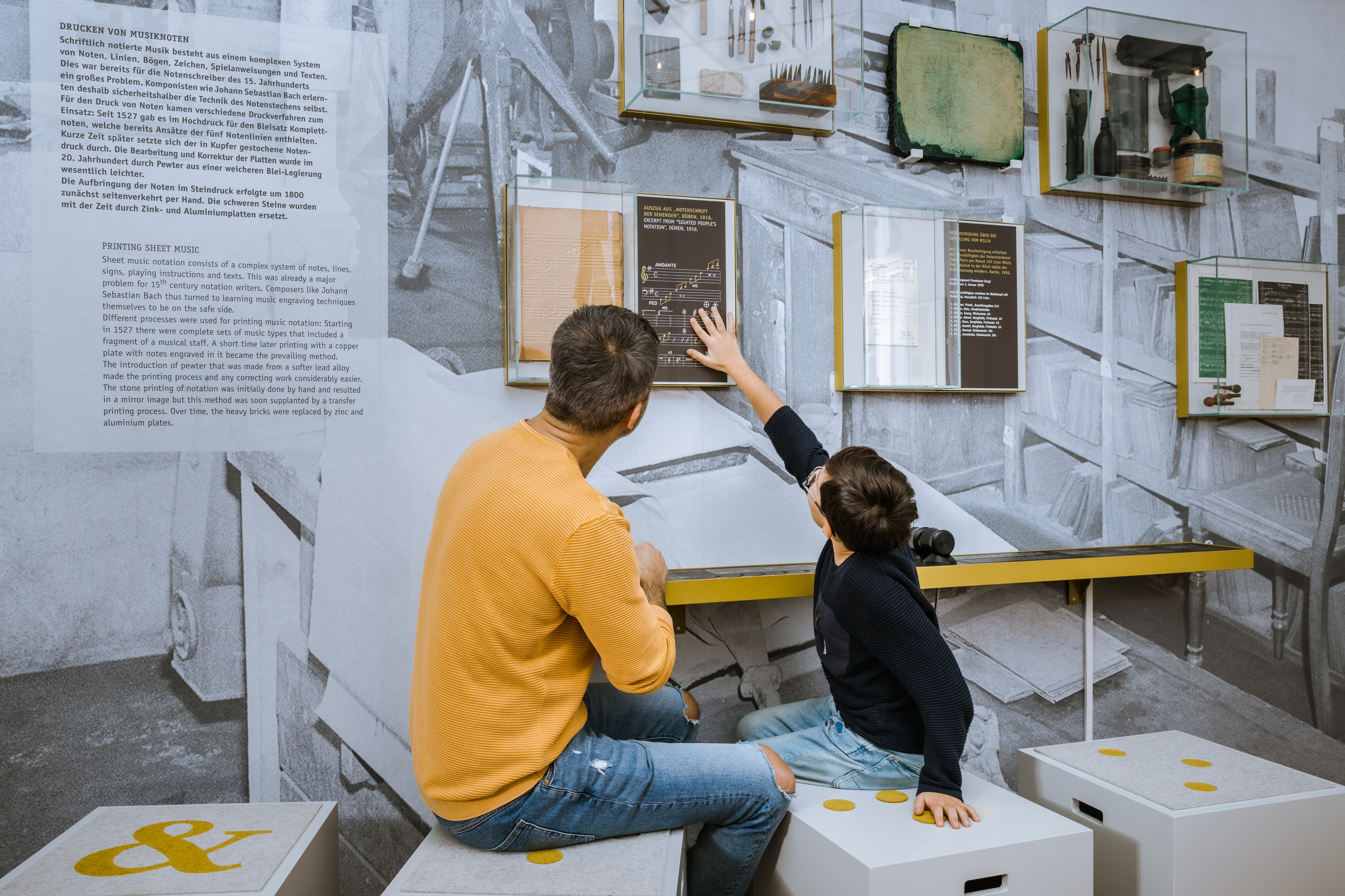 Ein Mann und ein Junge sitzen vor einer Ausstellungswand mit gedruckten Noten. An der Wand ist auch ein Tastobjekt, das der kleine Junge berührt.