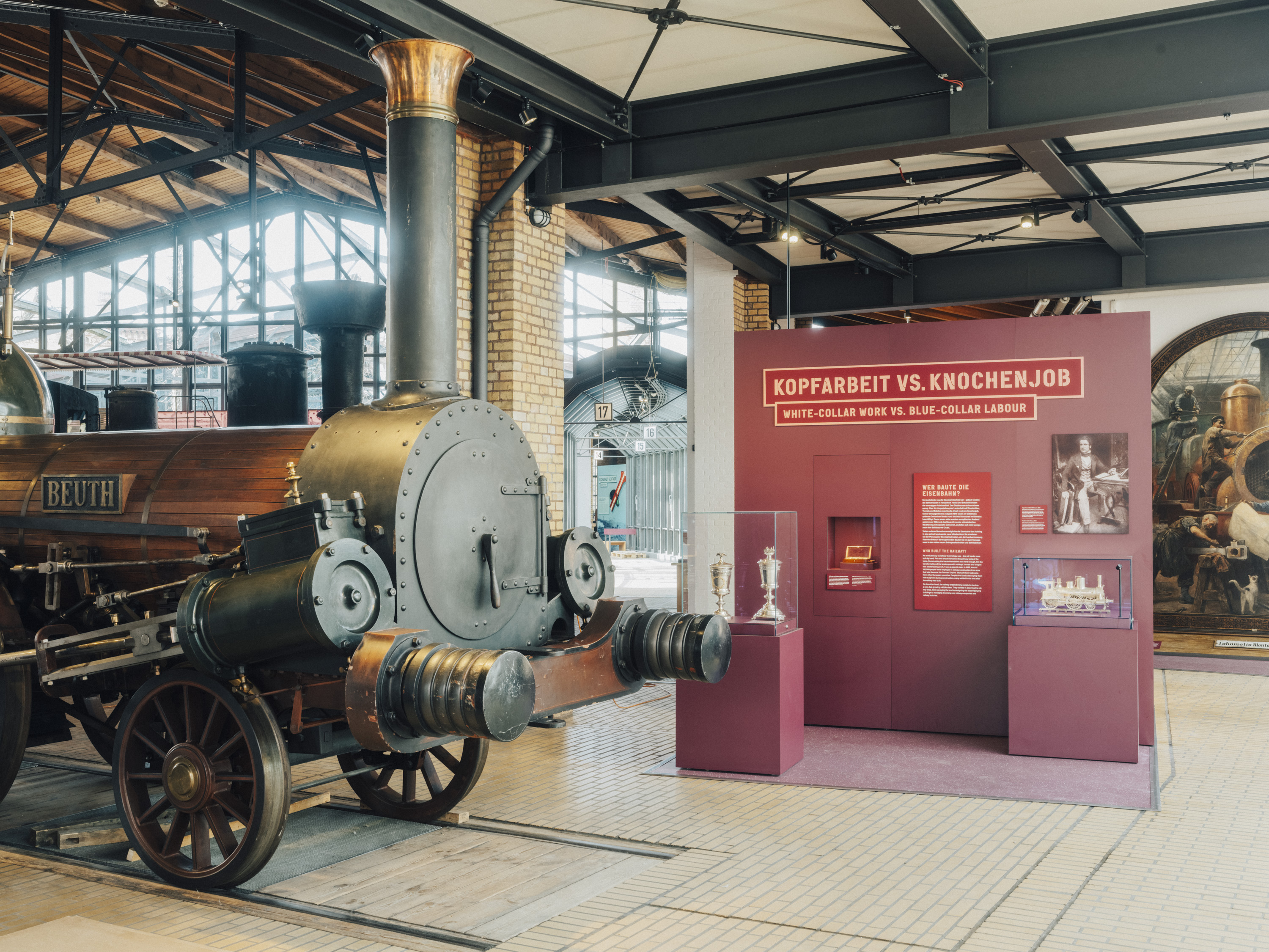Eine Lokomotive mit der Aufschrift „Beuth“ steht auf Gleisen in einem Lokschuppen. Eine weinrote Ausstellungsinsel trägt den Titel „Kopfarbeit vs. Knochenjob“.