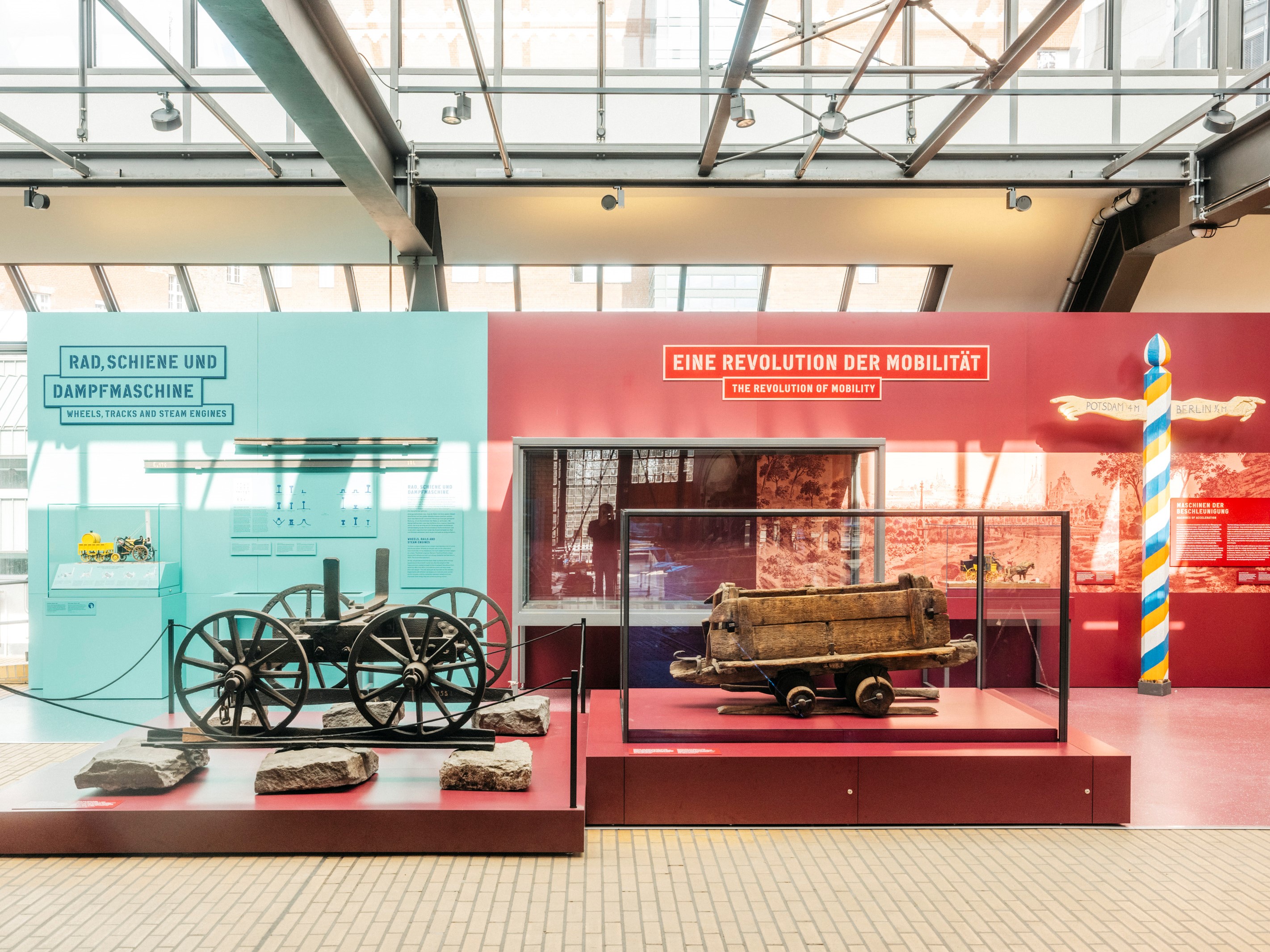 Rechts im Bild ist eine kulturgeschichtlich orientierte Ausstellungseinheit der Ausstellung Schienenverkehr mit der Überschrift „Eine Revolution der Mobilität“ auf einer weinroten Fläche zu sehen. Links wird eine Ausstellungseinheit mit der Überschrift „Rad, Schiene und Dampfmaschine“ vor einer türkisfarbenen Fläche gezeigt, die technische Prinzipien erklärt. Im Vordergrund zwei Objekte in Vitrinen.