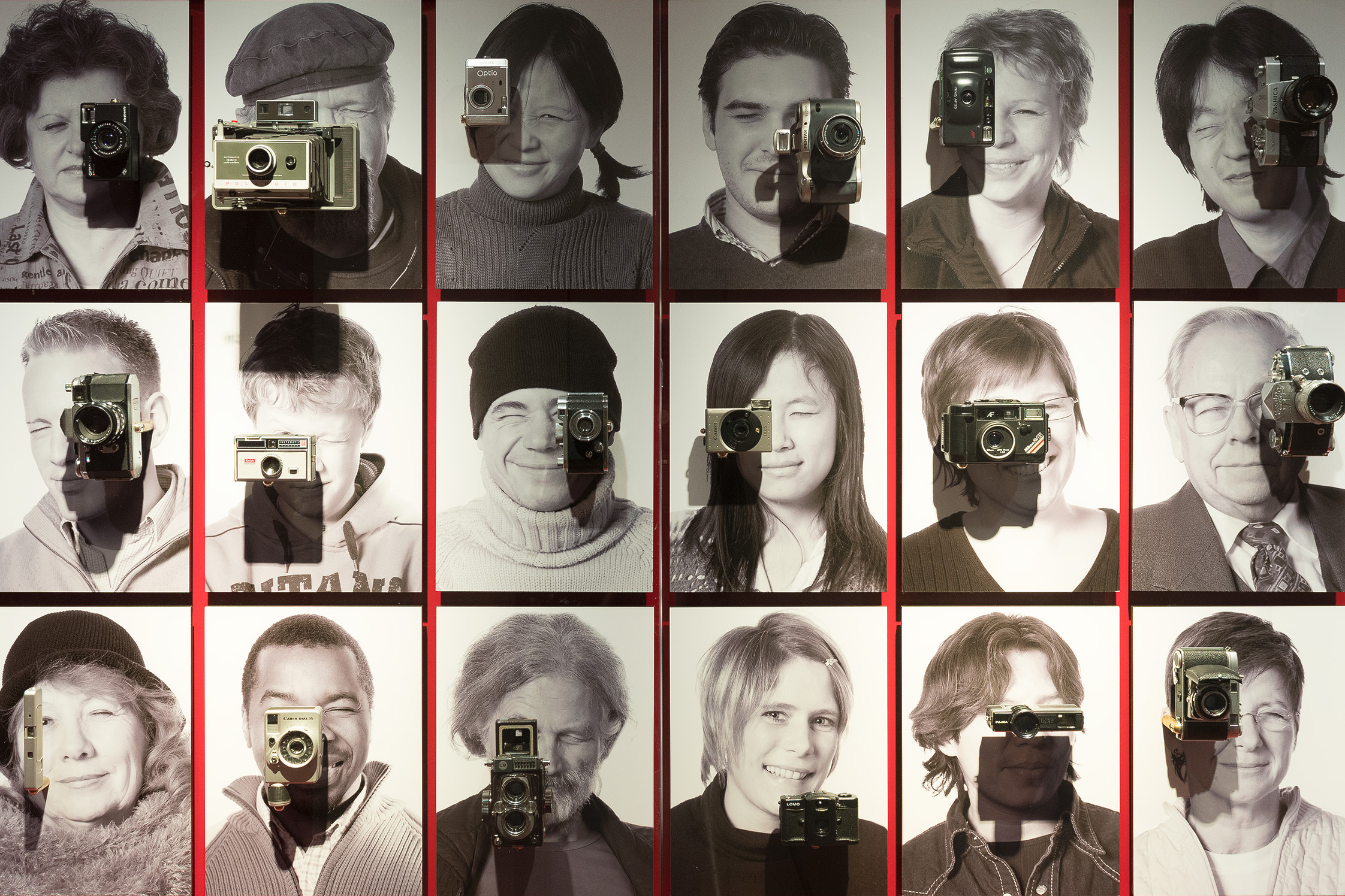 18 Schwarzweiß-Porträts von Männern und Frauen unterschiedlichen Alters sind in drei Reihen zusammengestellt. Vor ihnen ist jeweils eine historische Fotokamera angebracht. Es wirkt, als würden sie damit fotografieren.
