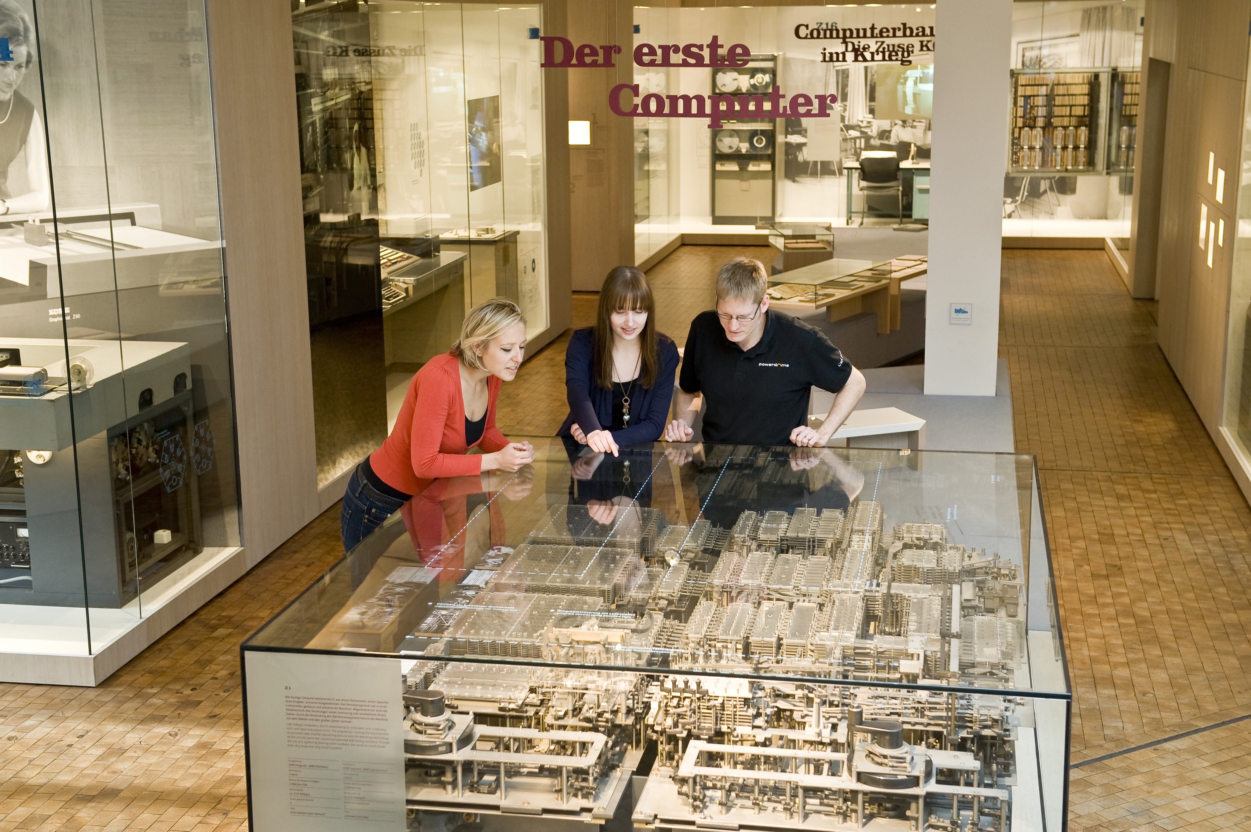 Zwei Frauen und ein Mann beugen sich über eine große Glasvitrine, in der das mechanische Rechenwerk der Z1 zu sehen ist.