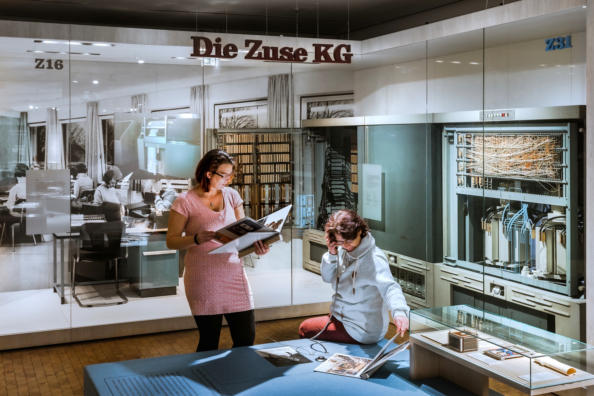 Blick in die Informatik-Ausstellung: Eine Frau sitzt auf einem Podest und hält sich den Hörer einer Hörstation ans Ohr. Eine andere Frau steht neben ihr und liest in einem großen Heft. In wandhohen Vitrinen im Hintergrund sind historische Rechenmaschinen zu sehen. Von der Decke hängt der Schriftzug „Die Zuse KG“. 