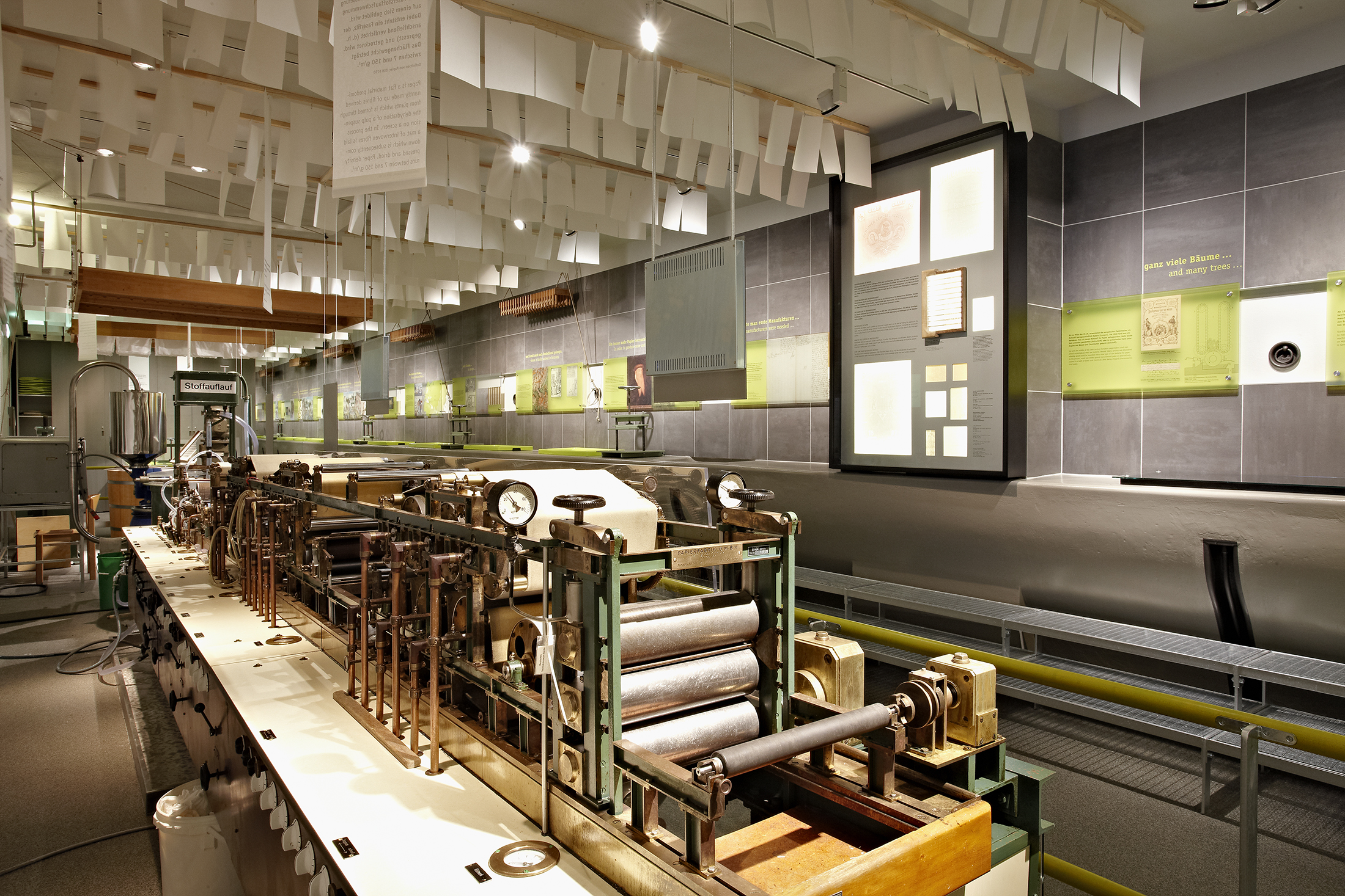 Blick in die Ausstellung Papiertechnik. Im Vordergrund steht eine mehrere Meter lange Maschine, von der Decke hängen Papierbögen, an der Wand sind Bilder und Texte angebracht.