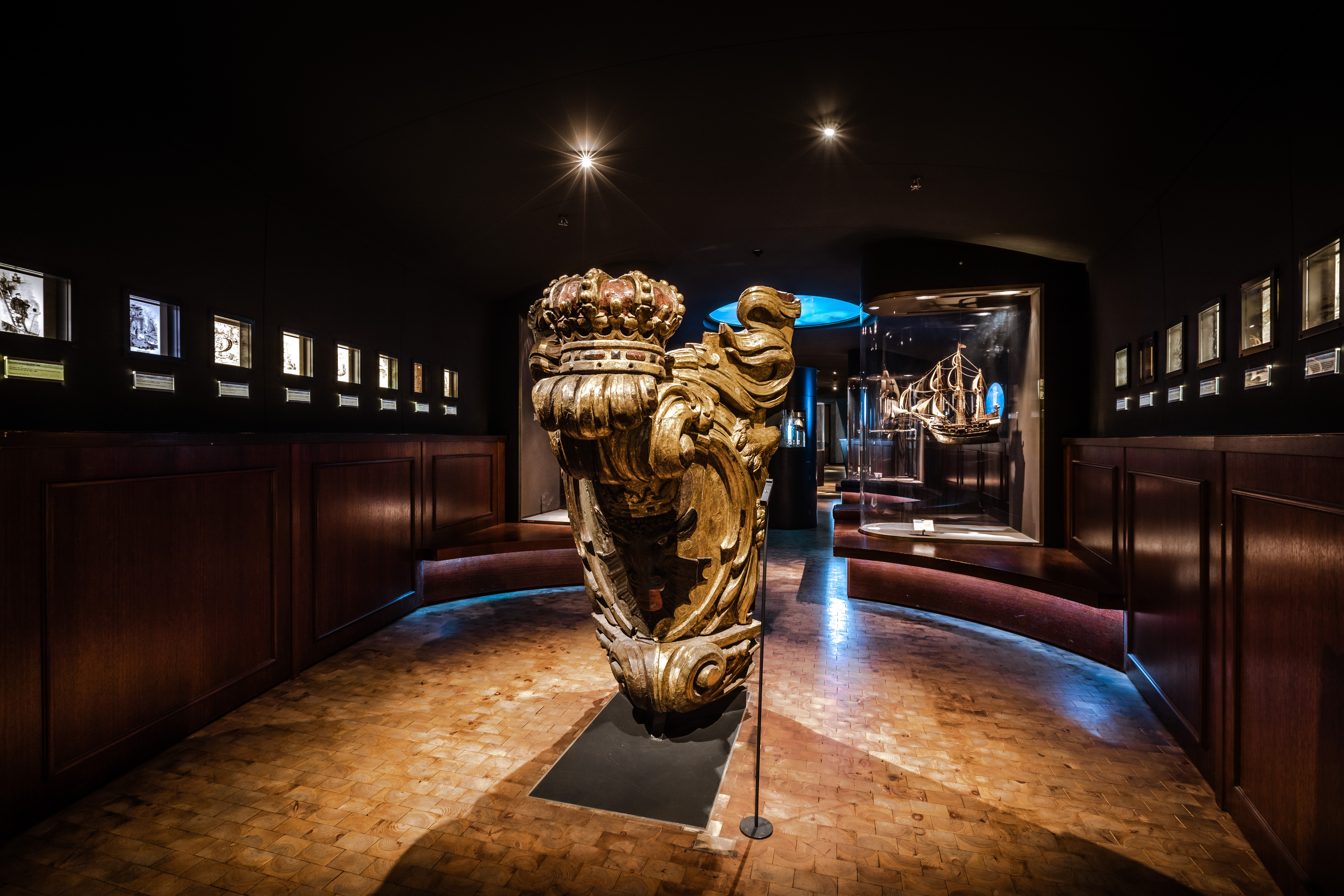 Blick in die Schifffahrt-Ausstellung: Zentral im Raum steht eine hölzerne Galionsfigur. An beiden Seiten verläuft ein Band aus kleinen Leuchtkästen. Im Hintergrund ist eine Vitrine mit einem Schiffsmodell zu sehen.