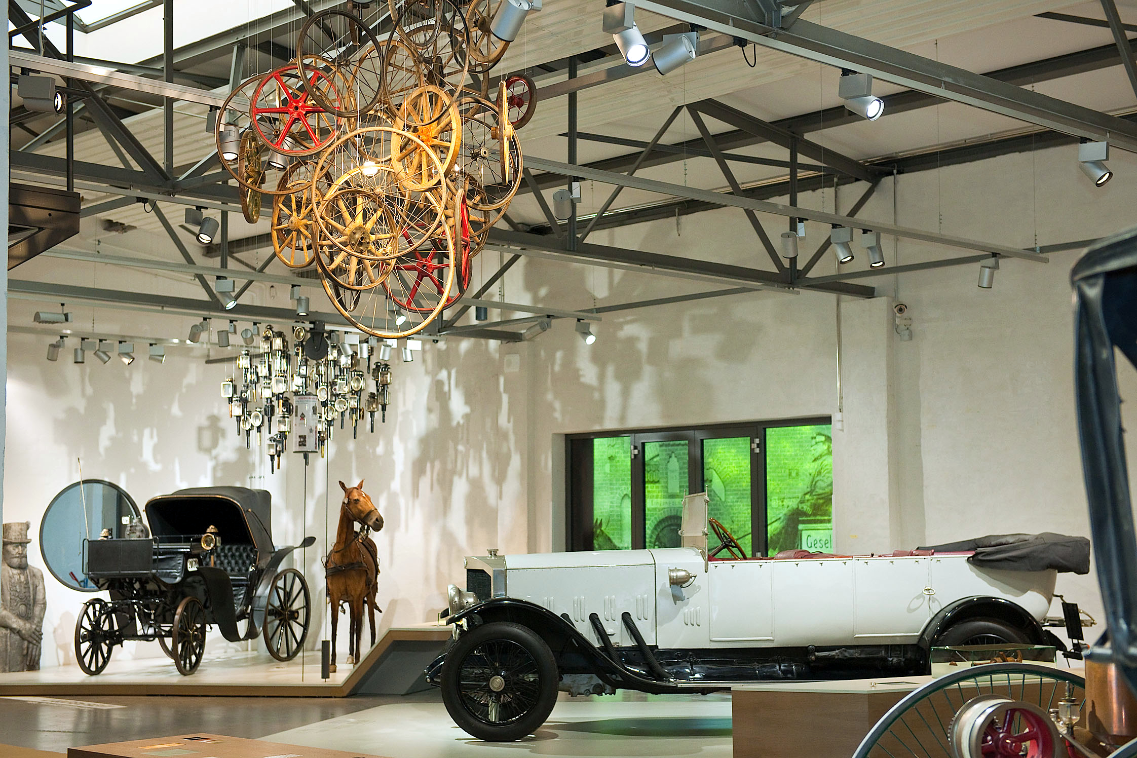 Blick in die Straßenverkehr-Ausstellung: Ein weißes historisches Cabriolet, eine Kutsche mit Pferd und eine Deckeninstallation aus verschiedenen Holz-Rädern.