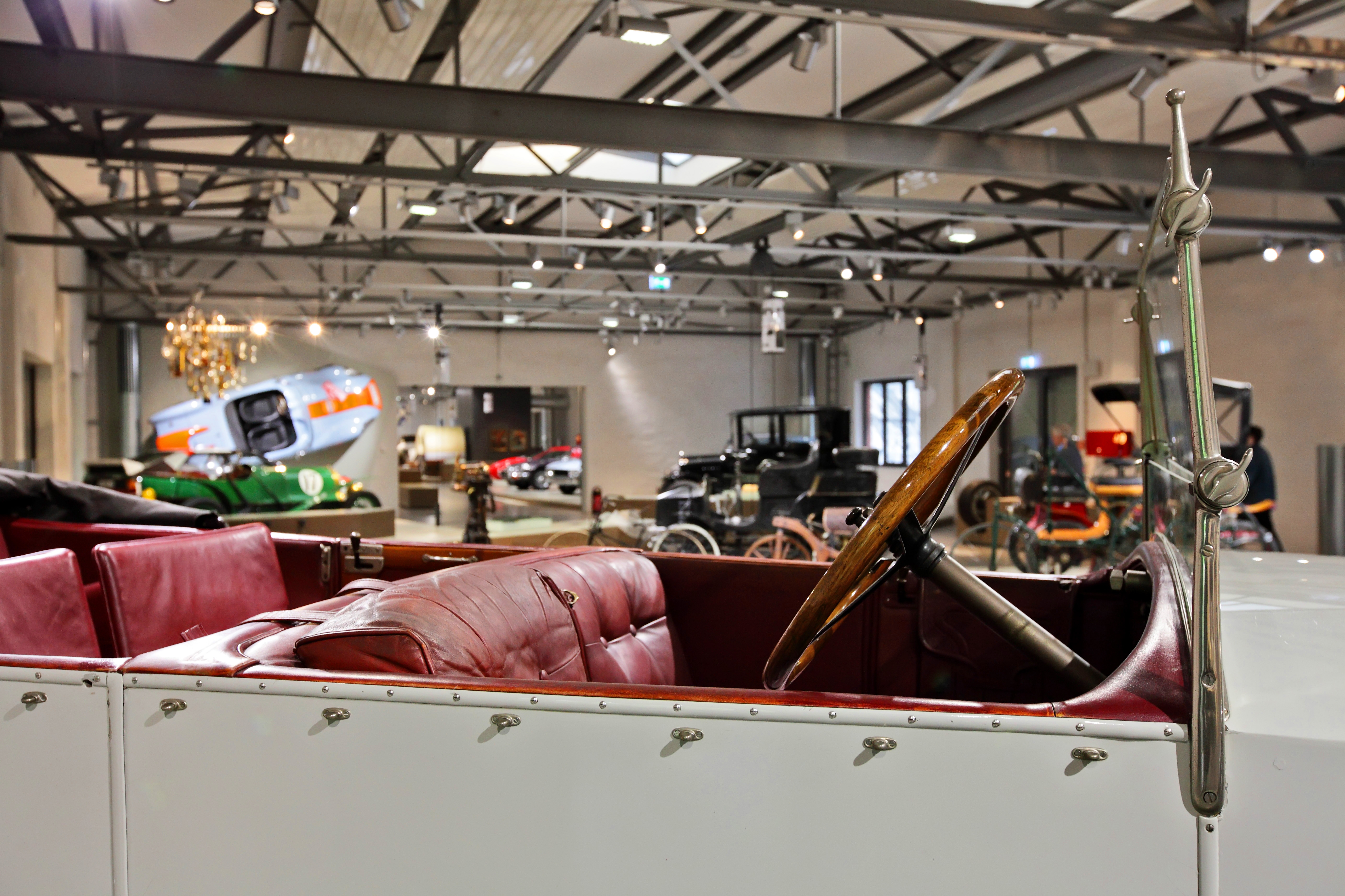 Blick über das offene Verdeck eines historischen weißen Cabriolets in die Straßenverkehr-Ausstellung. Sitze und Verkleidung sind mit dunkelrotem Leder bezogen, das Lenkrad ist aus dunklem Holz. 
