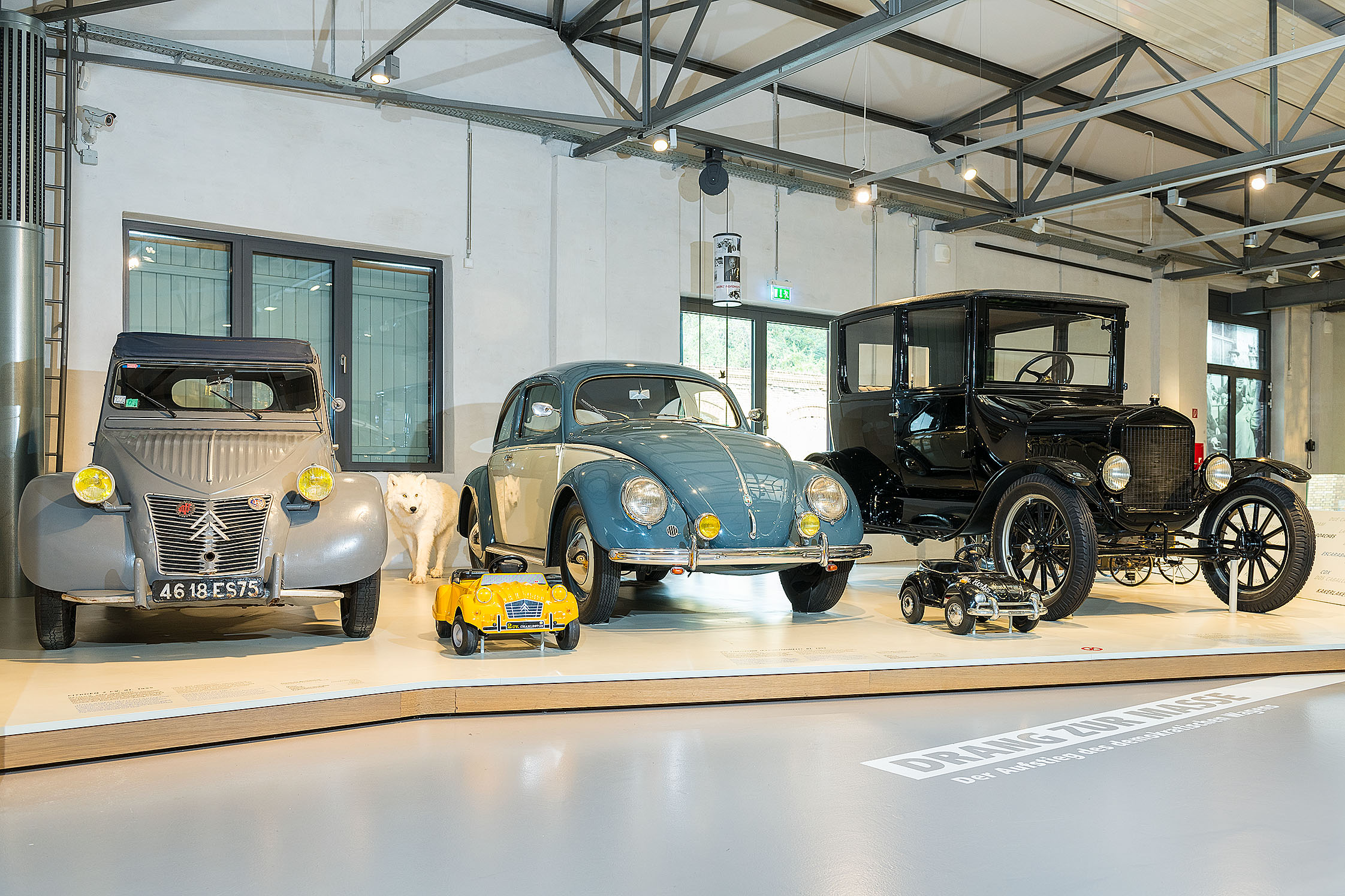 Auf einem niedrigen Podest stehen drei Automobile. Von links nach rechts: eine graue Citroën Ente, ein blauweißer VW Käfer und ein schwarzer historischer Wagen.