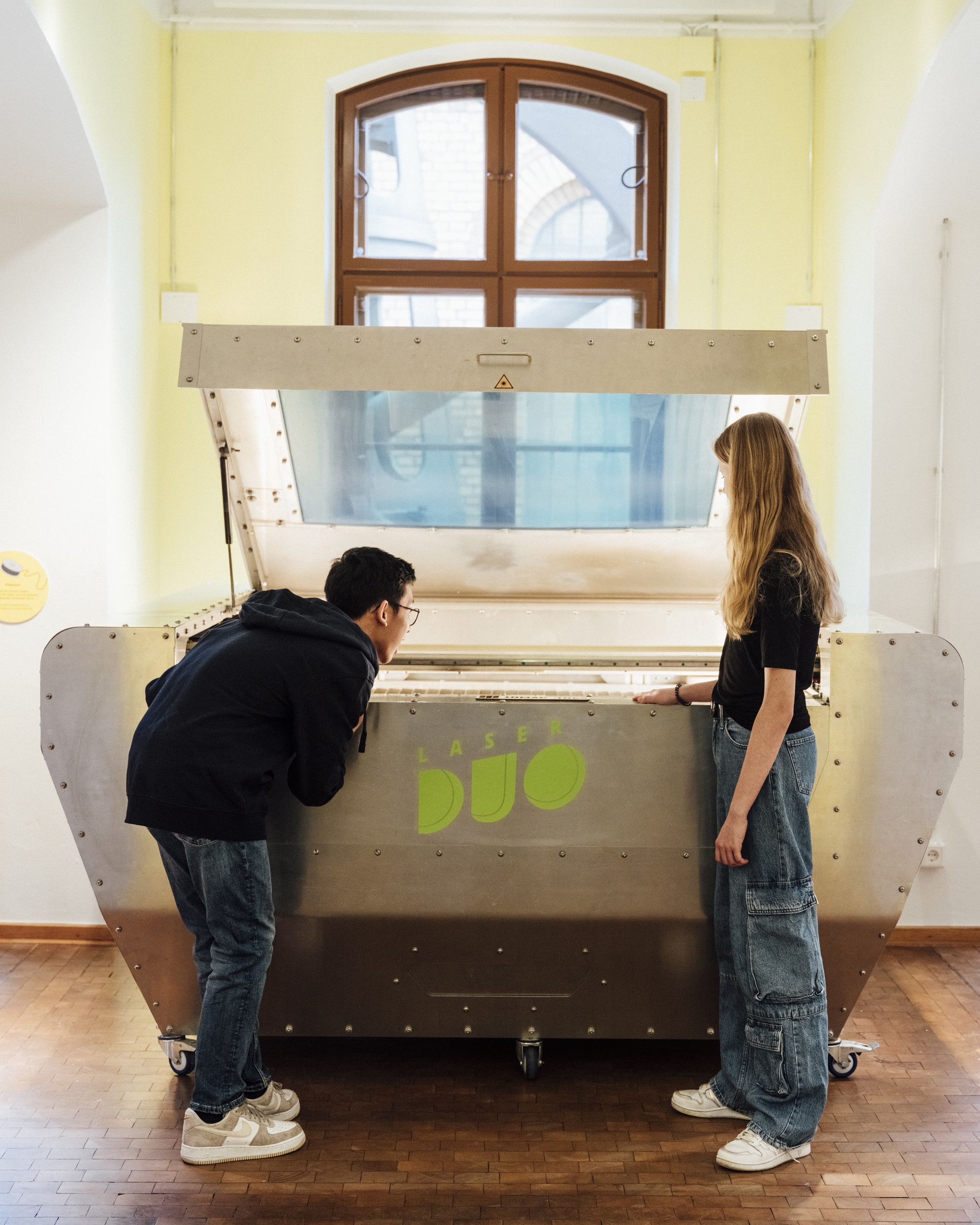 Ein Mädchen und ein Junge betrachten ein großes, silberfarbenes technisches Gerät. Auf dem erät steht „LaserDuo“. Der Junge beugte sich über die Öffnung des Geräts, das Mädchen steht neben ihm und betrachtet das Innere des Geräts.