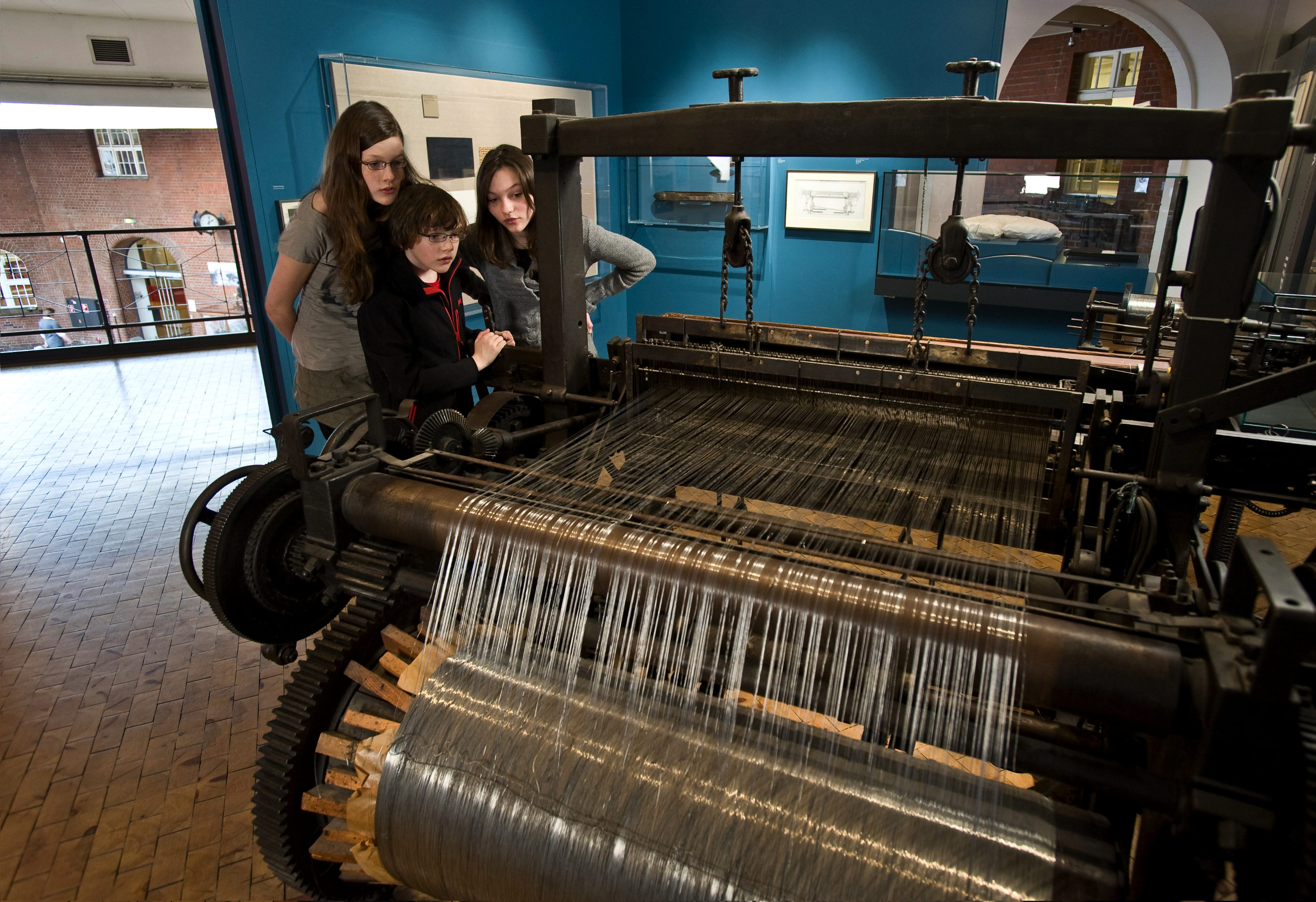 Drei Kinder betrachten einen großen, historischen Drahtwebstuhl in der Ausstellung. 