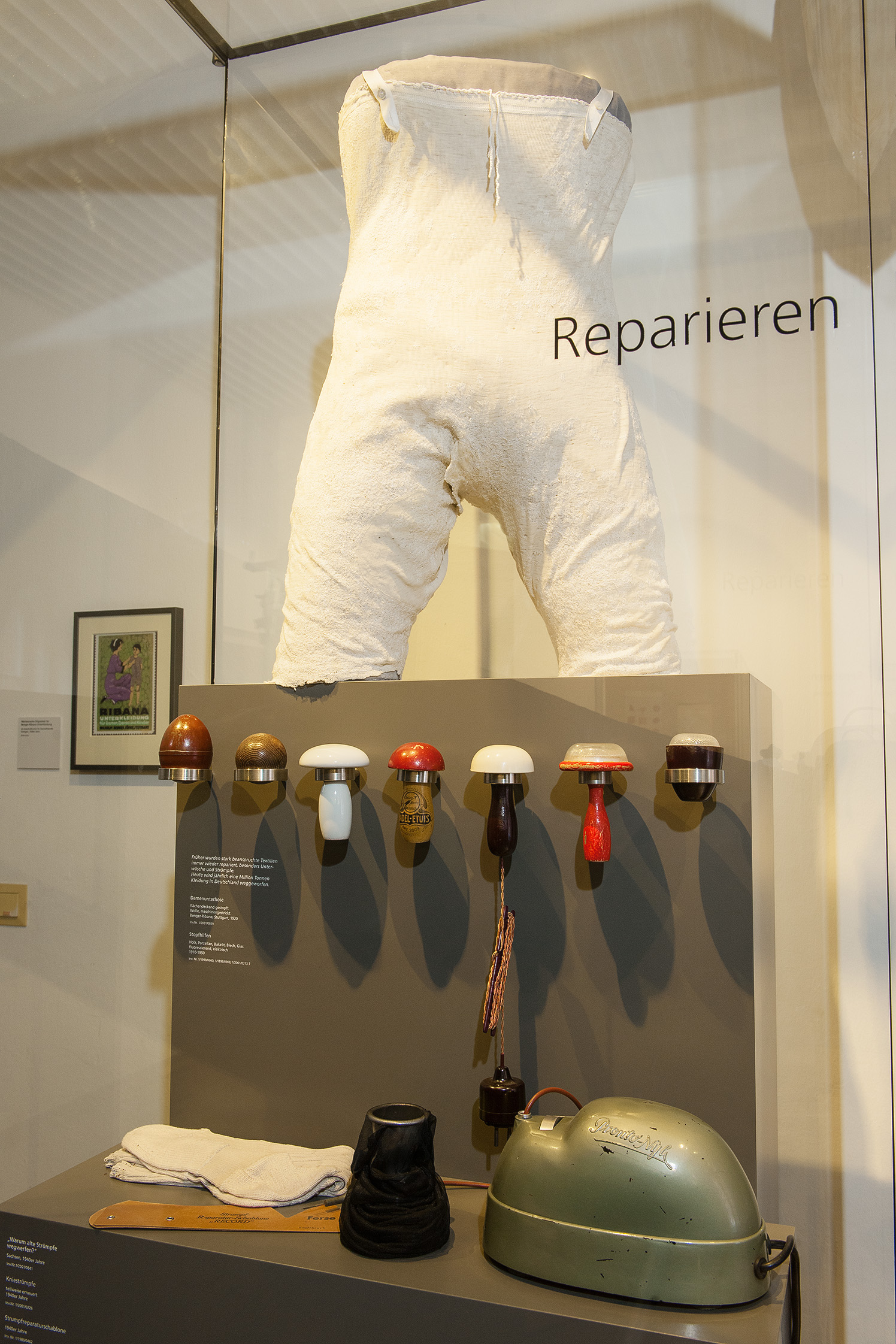 Eine vielfach gestopfte, weiße Unterhose auf einem Sockel in einer Vitrine. Unterhalb des Sockels sind verschiedene Stopfhilfen, die Pilzen ähneln, nebeneinander aufgereiht.