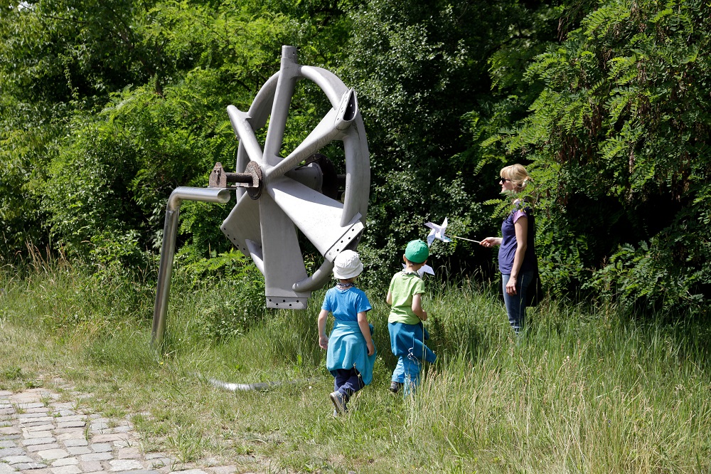 Zwei Kinder und eine Frau betrachten eine Windrad-Nabe, die an einem Metallgestell befestigt ist, das in hohem Gras steht.