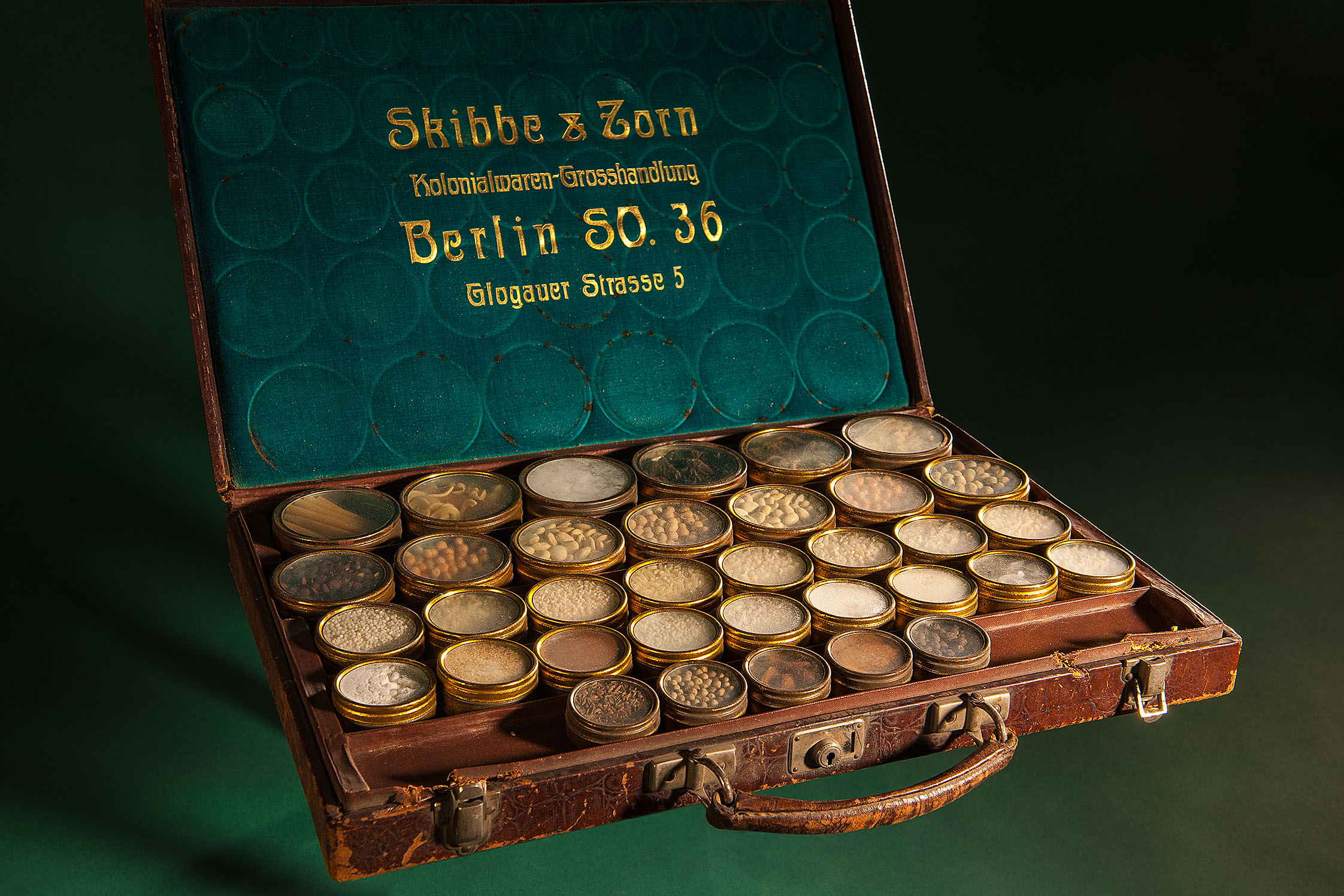 Lederkoffer mit verschiedenen kleinen Gefäßen, in denen sich Proben kolonialer Produkte wie Kakao befinden. Die Innenseite des Kofferdeckels trägt den Firmennamen Skibbe & Zorn.