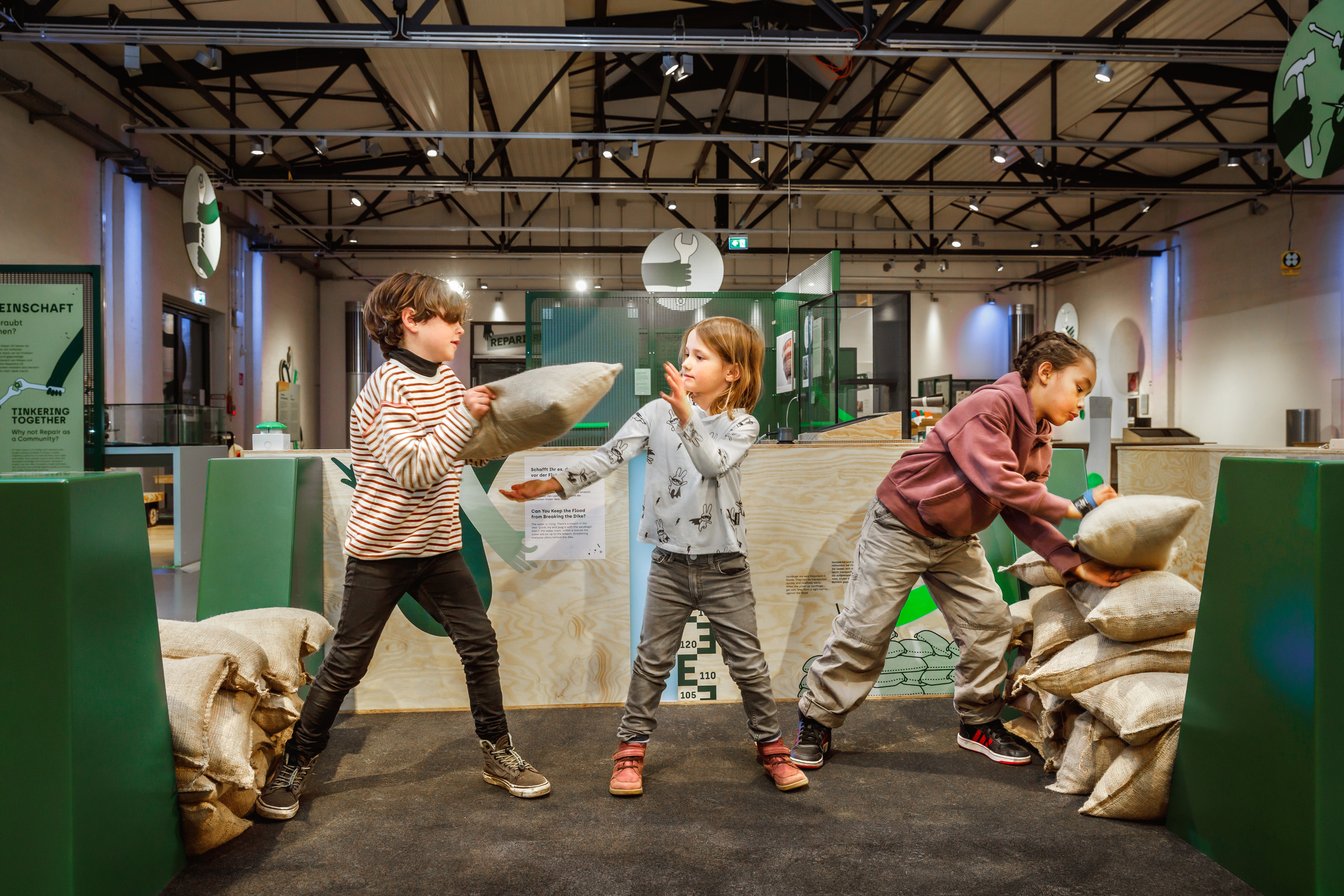 Drei Kinder reparieren in einer Ausstellungshalle einen Spielzeug-Deich aus Sandsäcken. Sie bilden eine Kette und reichen sich die Säcke, um eine Lücke im Deich zu flicken.
