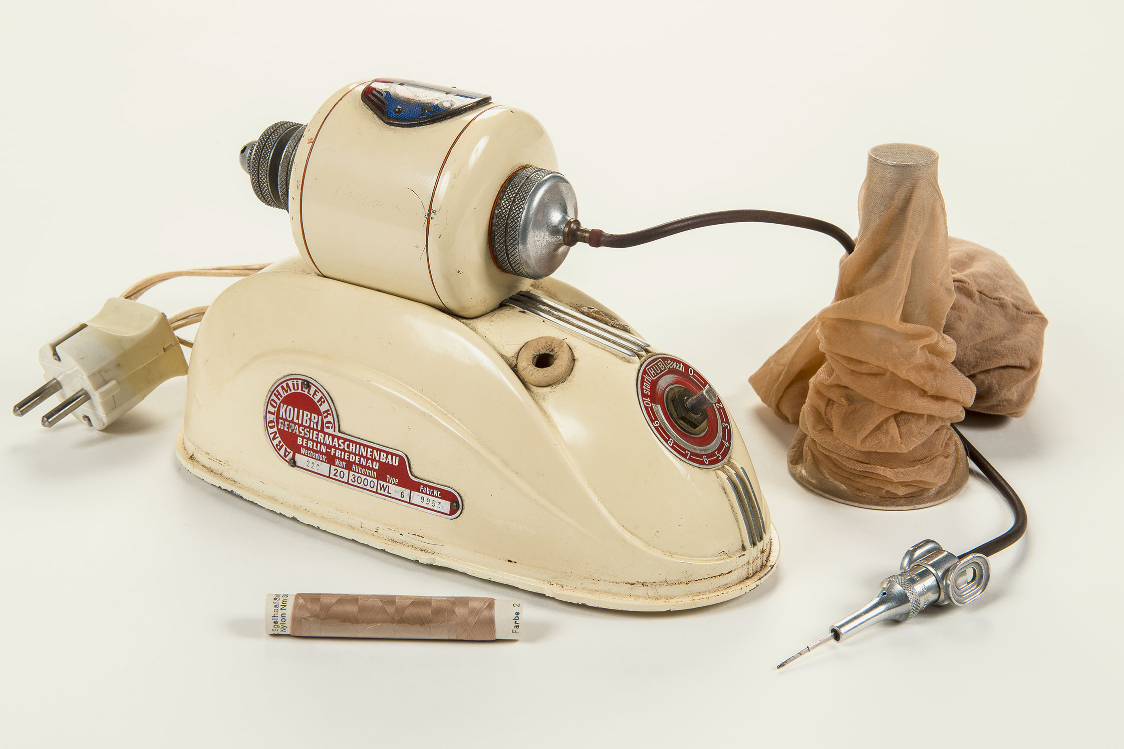 Zu sehen ist eine cremefarbige Maschine, die entfernt an ein Bügeleisen erinnert. Sie hat ein breites Standbein und einen Stecker, einen braunen Schlauch und eine silberfarbene Nadel. Daneben steht ein Ständer mit einer beige-braunen Feinstrumpfhose, davor liegt eine Garnrolle in der gleichen Farbe.