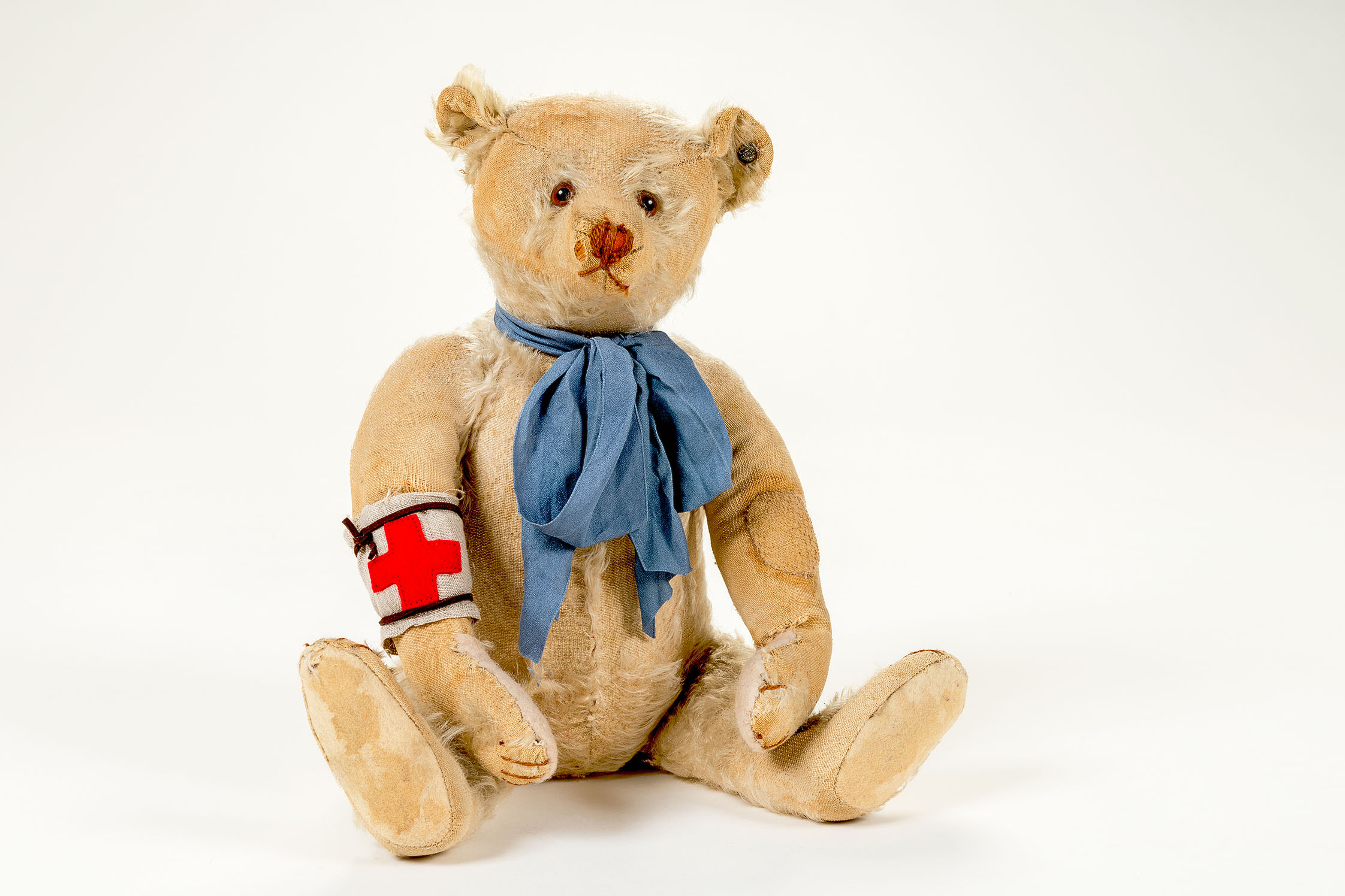 Zu sehen ist ein beiger Teddybär mit offensichtlich kaputten und abgewetzten Stellen. Er trägt eine blaue Schleife um den Hals und eine weiße Armbinde mit rotem Kreuz.