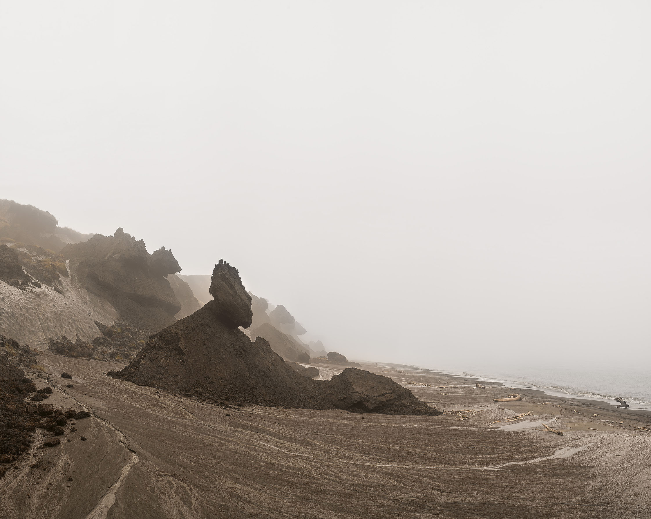 An einem Strand mit braunem Sand ragen dunkle Erdhügel in Richtung des Meeres. Sie sehen aus wie Eisberge auf dem Trockenen. Himmel und Meer sind gleichmäßig grau. Ein Horizont ist nicht zu erkennen. 