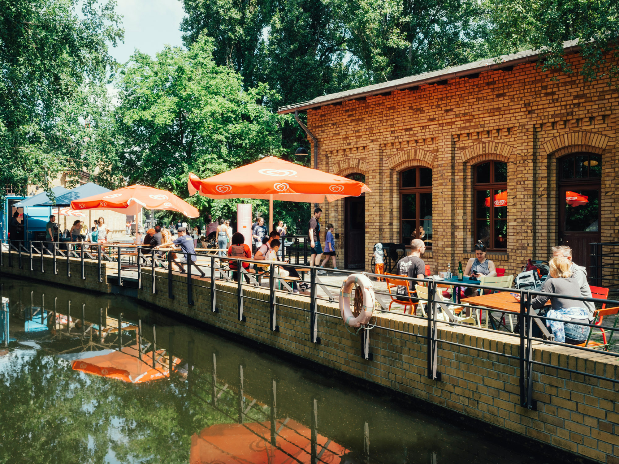 Blick auf den Museumsteich und ein Backsteingebäude, am Ufer sind bunte Gastronomie-Schirme, Tische und Bänke zu sehen, an denen Menschen sitzen. Im Hintergrund ein blauer Food-Container.